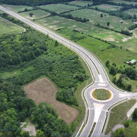 An aerial view of a roundabout intersection and 4-lane roadway through forest and farmland.