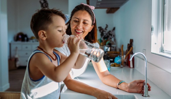 A young boy drinking a glass of water beside his mother.